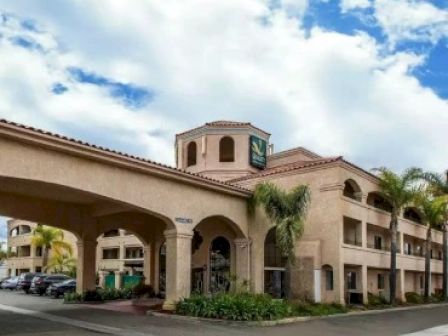 The image shows a multi-story hotel building with an archway entrance, palm trees, and parked cars, set against a cloudy blue sky.