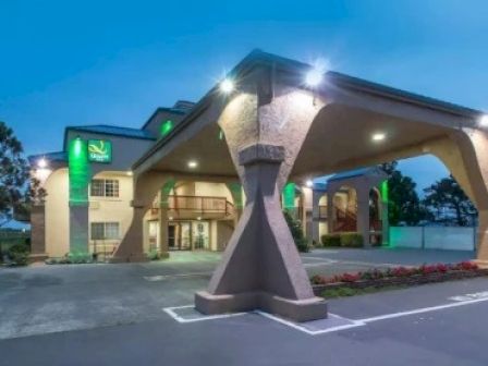 The image shows the exterior of a hotel with a covered entrance and multiple floors, illuminated by exterior lights at dusk.