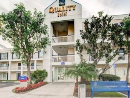 The image shows the exterior of a Quality Inn hotel, featuring trees, a blue bench, and a manicured garden at the entrance.