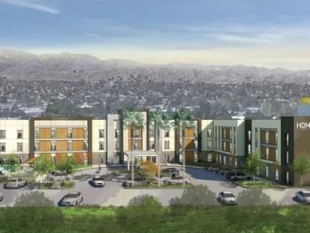 This image shows a hotel building with multiple stories, a parking lot in front, and a mountainous background under a partially cloudy sky.