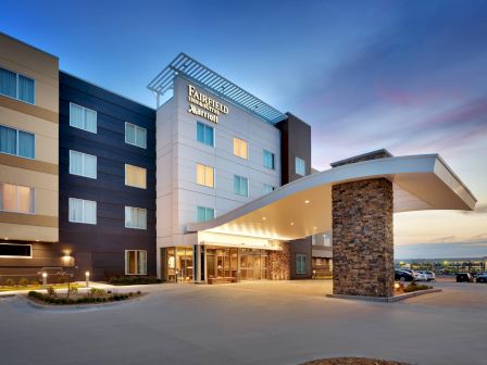 The image shows the modern exterior of a Fairfield Inn & Suites hotel at dusk, with a well-lit entrance and a clear evening sky.