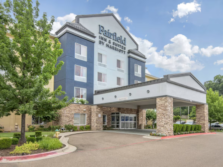 The image shows the exterior of a Fairfield Inn & Suites hotel with a parking area and landscaped surroundings under a partly cloudy sky.