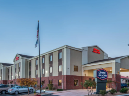 The image shows the exterior of a Hampton Inn & Suites hotel building with an American flag, parked cars, trees, and a clear sky in the background.