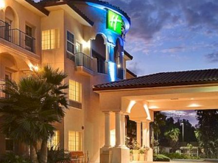 The image shows the entrance of a hotel building with illuminated signs, palm trees, and a well-lit exterior in the evening.