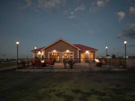 A well-lit house with a red roof and a fenced patio with chairs and fire pits, set against a dusky sky with scattered clouds.