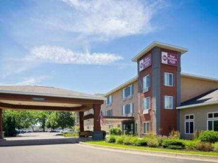 The image shows the exterior of a modern hotel building with a covered entrance, clear blue sky, and well-maintained landscaping.