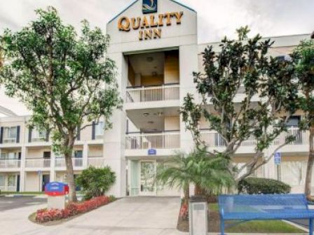 This image shows the exterior of a Quality Inn hotel, featuring a well-maintained walkway, trees, a bench, and a blue and red sign.