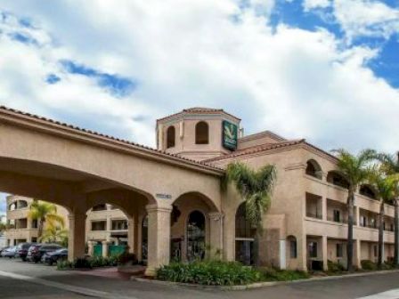 The image shows a Mediterranean-style hotel building with arches, palm trees, and a cloudy blue sky above. There are cars parked to the left.