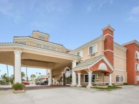 The image shows the exterior of a hotel with a large entrance canopy supported by pillars, a beige and red color scheme, and clear skies above.