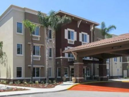 The image shows a modern, multi-story building with palm trees and a covered entrance, likely an apartment complex or hotel.