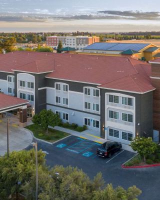 An aerial view of a three-story building with red roofs, parking spaces, and surrounding greenery, taken during the day.