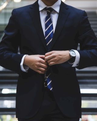 A person in a business suit adjusts their jacket while standing in front of a staircase, with a modern interior setting in the background.