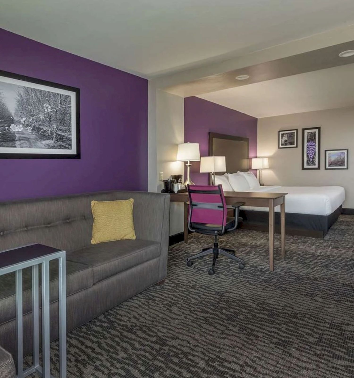 A hotel room with a purple accent wall, a couch, a desk with a chair, a bed, framed art, and ample lighting.