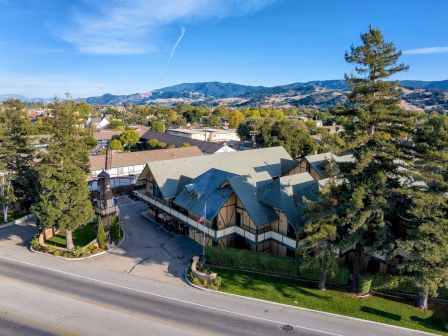 Aerial view of a building with a green roof and a European architectural style, surrounded by trees and mountains in the background.