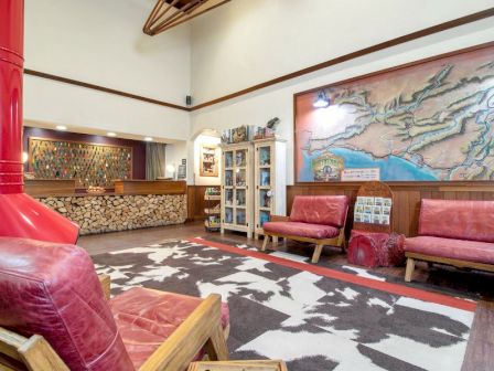 A cozy room with red leather chairs, a patterned rug, a wooden shelf, a wall map, and a wood-burning stove with stacked firewood.