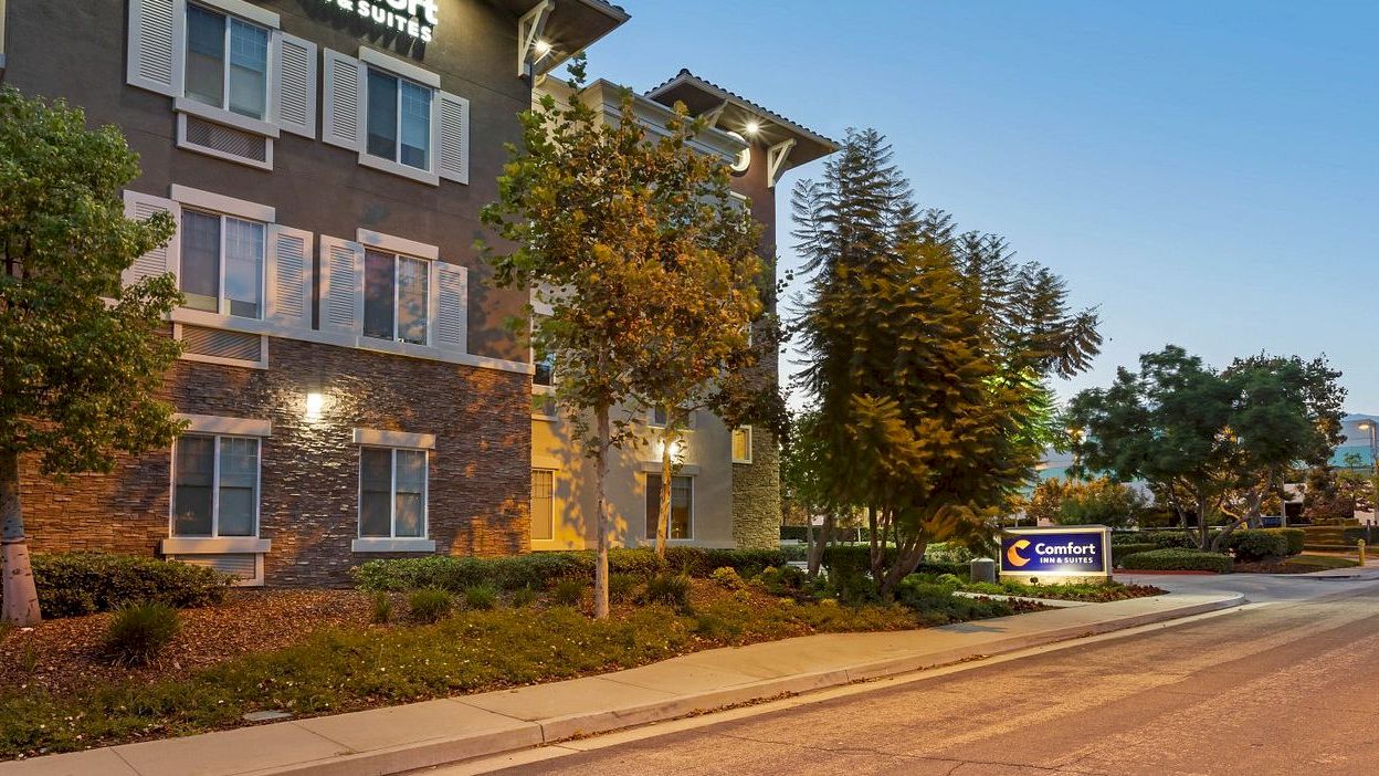 A hotel building with exterior lighting, trees, and a "Comfort" sign next to the sidewalk in a quiet neighborhood.