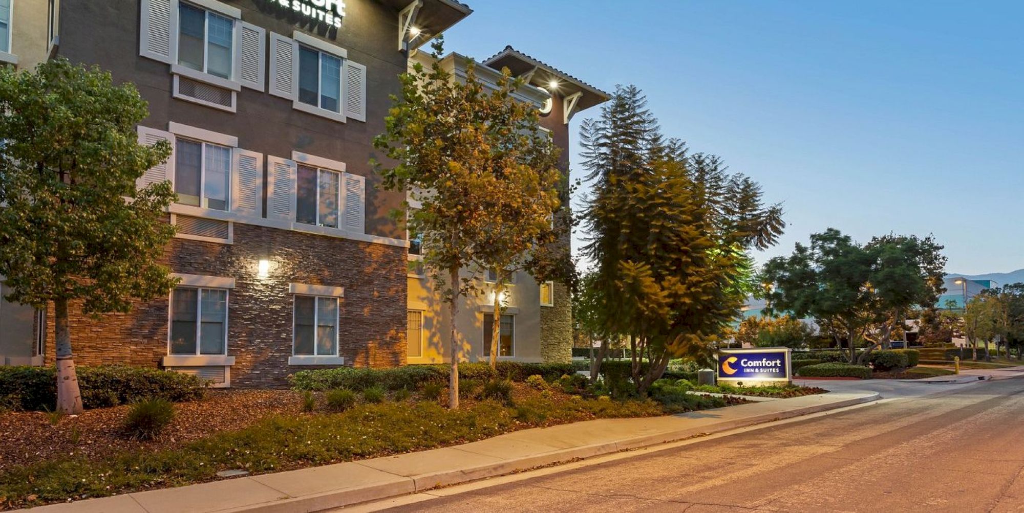 A hotel building with exterior lighting, trees, and a "Comfort" sign next to the sidewalk in a quiet neighborhood.