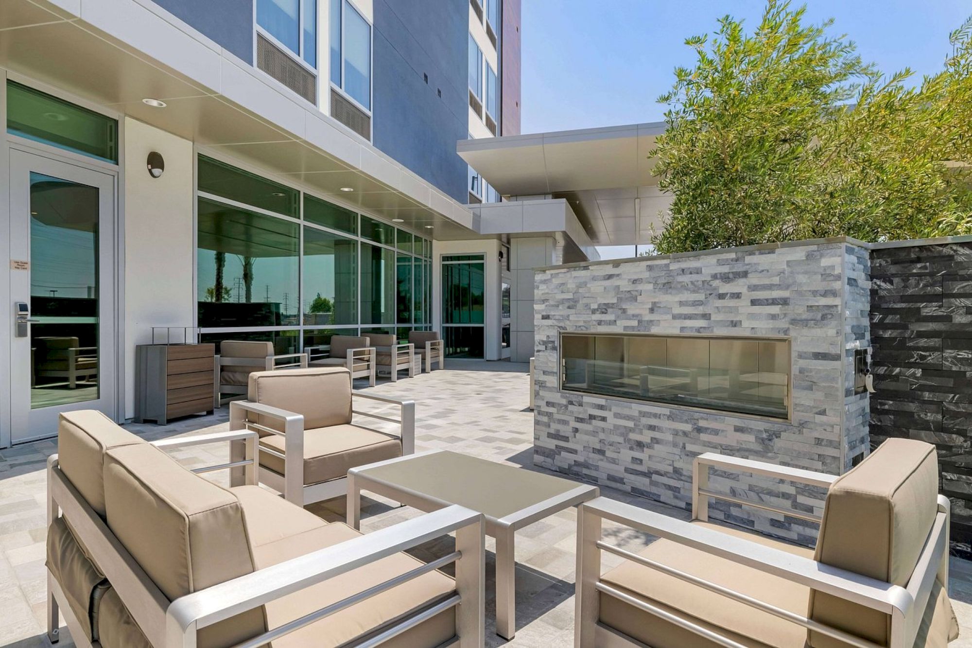 The image shows an outdoor seating area with beige cushioned chairs, a table, and a built-in modern fireplace against a stone wall.