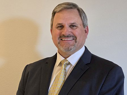 The image shows a man with short gray hair, wearing a dark suit, white shirt, and yellow tie, smiling while standing against a plain background.