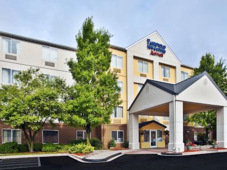The image shows the exterior of a hotel building with a prominent entrance and several windows, surrounded by trees and greenery.