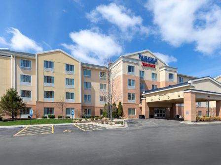 This image shows a four-story hotel building with a parking area in front, featuring a blue sky and a few scattered clouds.