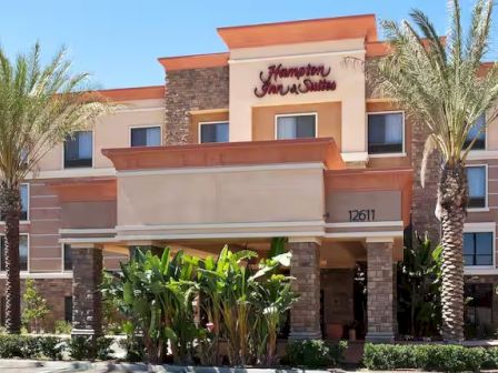 The image shows a Hampton Inn & Suites hotel entrance surrounded by palm trees and plants under a clear blue sky.