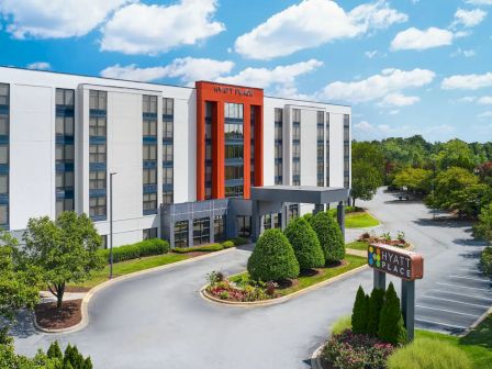 The image shows a modern hotel building with a sign for Hyatt Place, surrounded by landscaped greenery and a parking area.