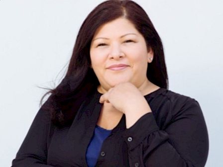 The image shows a person with long hair wearing a blue top and black jacket, smiling with arms crossed, set against a plain light background.