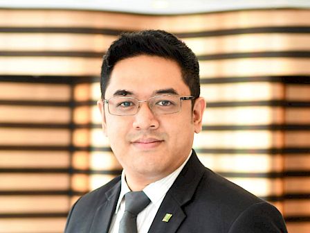A man in a suit and tie, with glasses, stands against a background with horizontal stripes, smiling professionally.
