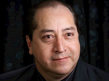 A person is wearing a dark suit with a light gold tie, set against a dark background.