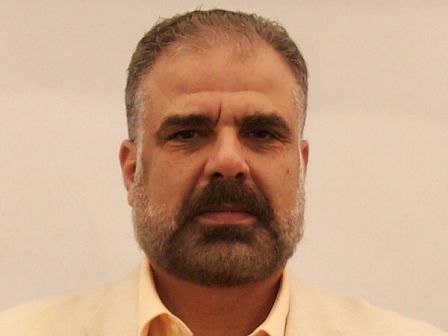 The image shows a person with a beard wearing a light-colored blazer and a shirt, against a plain background.