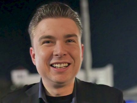 A smiling man in a dark blazer and blue shirt poses for the camera at night, with a blurred background and warm lighting.