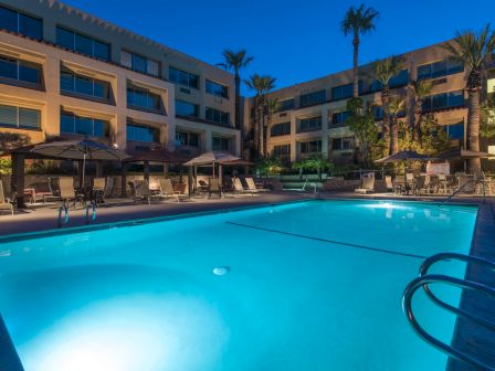 A bright outdoor hotel pool at dusk, surrounded by palm trees, lounge chairs, and a three-story beige building with balconies.