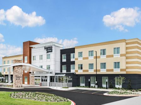 A modern hotel or motel with a curved entrance, beige and gray exterior, multiple windows, a porte-coch&egrave;re, and a manicured driveway and lawn.