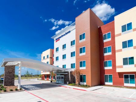 A modern multi-story hotel or apartment building with a curved covered entrance, brick and beige facade, blue sky, and neat landscaping.