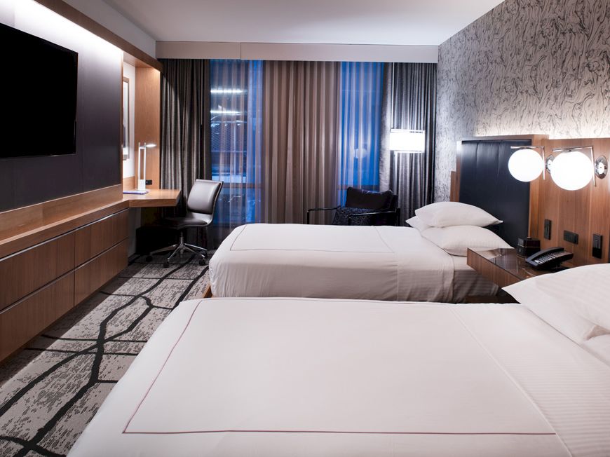 A modern hotel room with two beds, a TV, a desk with a chair, and a patterned wall. The room features contemporary decor and lighting.