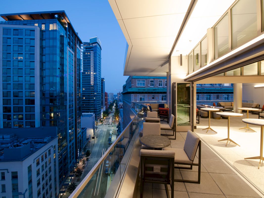 The image shows a modern cityscape with tall buildings and a rooftop patio furnished with tables and chairs at dusk.