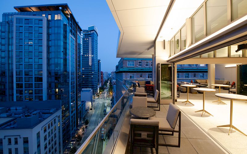 A modern cityscape at dusk with tall buildings, viewed from a stylish balcony featuring seating and tables, overlooking a street below.
